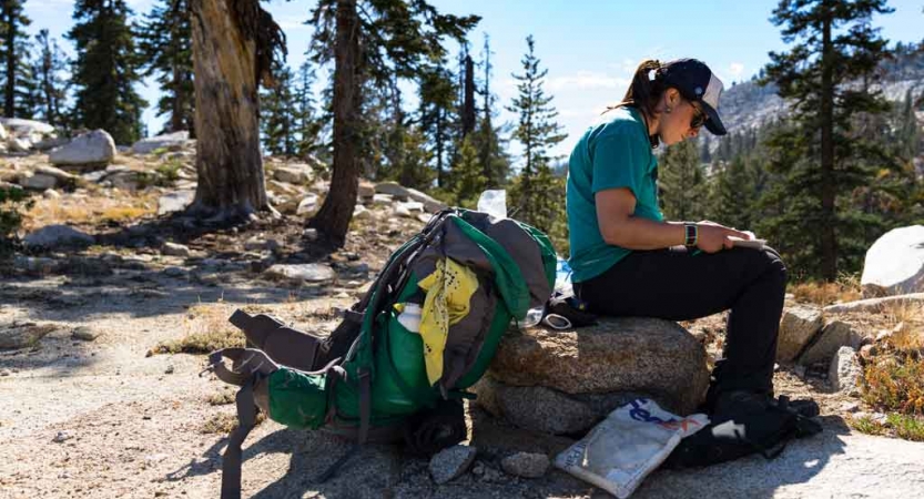 A person sits on a rock in with their backpack resting on the ground. They appear to be reading or journaling. 